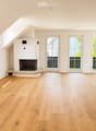 Bright, unfurnished room with wooden flooring, white walls, and a fireplace. Three large windows and doors lead to an outdoor area.