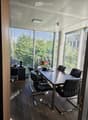 Modern office space with large windows, wooden floor, black leather chairs, a long table, a small cabinet, and a potted plant.