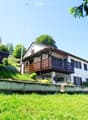 Two-story house with a raised ground floor, white exterior, wooden balcony, and surrounded by greenery.
