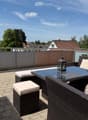 A modern rooftop terrace with a wicker seating set, glass table, and lantern, overlooking a residential area with trees and houses.
