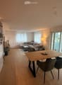 Open-plan living and dining area with light wood flooring, white walls, large windows, and a sliding glass door leading to a balcony. Features a grey sectional sofa, coffee table, dining table with chairs, and a kitchen counter with a vase and plant.
