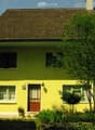 Single-story yellow house with a brown tiled roof, white-framed windows, and a small front garden with trees and shrubs.