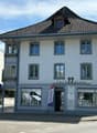 A three-story building with a classic European architectural style, featuring a tiled roof, multiple windows, and a storefront with signage reading "farm2kasse" and a RE/MAX logo.