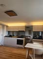 A modern, open-concept living and dining area with a kitchen. The space features a wooden floor, white walls, and large windows. A sofa is positioned near the window, a dining table with chairs is in the center, and a compact kitchen area is visible with