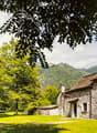 Stone house with a thatched roof, surrounded by lush green trees and a grassy field, with mountains in the background.