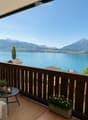 Balcony with wooden railing, overlooking a lake and mountains, furnished with a sofa, small table, candle, and potted plants.