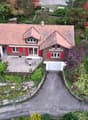A two-story house with a red-tiled roof and red shutters, surrounded by a well-maintained garden and driveway. The house features a garage and a small patio area with outdoor furniture.