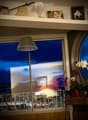 A cozy living space with large windows offering a city view, a floor lamp, a small table with a flower pot, and decorative items on the shelf above the window.