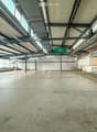 Large, empty industrial-style space with concrete floors, exposed metal beams, and large windows allowing natural light. Minimal furnishings, including a few stacked crates and a small table in the background.