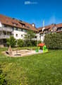 A well-maintained garden area with a small playhouse, benches, and a table, surrounded by a two-story residential building with balconies and a red-tiled roof.