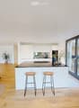 Open-plan living and dining area with a modern kitchen island, sofa, bar stools, and decorative vases. Large windows offer a view of the outdoor space.