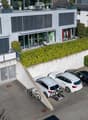 Modern multi-story residential building with large windows, balconies, and a parking area with several cars and bicycles.