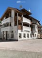 A multi-story building with white walls, wooden accents, and balconies. The building features a sign reading "PIZZERIA" and has multiple windows with curtains. The ground is paved with cobblestones.