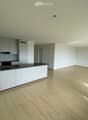 Empty room with light wooden flooring, white walls, and a modern kitchen area featuring grey cabinets, a black countertop, and stainless steel appliances.