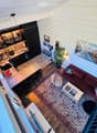 A modern, open-concept living and dining area with a red sofa, coffee table, round stool, and kitchen counter. The space features wooden flooring, patterned rugs, and a potted plant near the window.