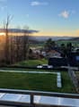 View from a balcony or terrace showing a grassy yard, surrounding houses, trees, and a scenic sunset view of hills and fields.
