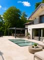 Modern outdoor patio with a swimming pool, white sofa, coffee table, dining area, and potted plants, connected to a house with a balcony and large windows.