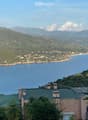 A scenic coastal view with a house on a hillside overlooking the sea, surrounded by lush greenery and distant mountains.