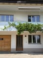 Two-story house with white exterior, wooden garage door, and balcony with green vines. Features multiple windows and a covered entrance area.