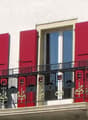 Balcony with black metal railing and decorative elements, red shutters, and large windows on a white building facade.