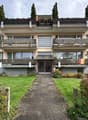 A three-story residential building with balconies, large windows, and a well-maintained garden in front.
