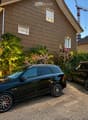Two-story house with wooden siding, multiple windows, and a well-maintained garden with various plants and flowers. Two black SUVs are parked in front of the house.