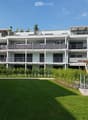 Multi-story apartment building with modern design, large balconies on each floor, green garden area in front, well-maintained surroundings.