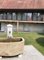 A two-story house with a balcony, stone walls, and a well-maintained garden featuring a stone water basin and a fountain.