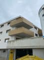 Modern multi-story building with white facade, large windows, and distinctive gold-colored balconies. Features a raised ground floor and a visible garage entrance.