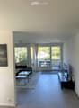 Modern living room with large windows and a balcony, featuring a sofa, coffee table, TV stand, and chairs.