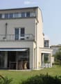 Modern two-story house with white exterior, large windows, balconies, and a covered patio area with outdoor furniture.