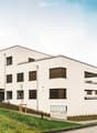 Modern multi-story residential building with white facade, multiple windows, balconies, and surrounding greenery.