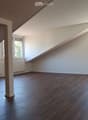 Empty attic room with wooden flooring, white walls, and large windows allowing natural light. The room features a slanted ceiling and a central support column.