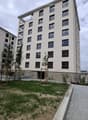 Modern multi-story residential building with white facade, multiple windows, and balconies. The ground level features a landscaped area with grass, small trees, and a paved walkway.