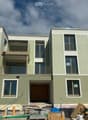 Modern three-story building with green and gray exterior, large windows, and balconies. Construction materials and equipment are visible in front.