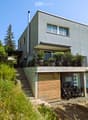 Modern two-story house with grey facade, large windows, balcony, and steps leading to a wooden garage area with bicycles parked nearby.