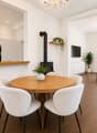 Open-plan living and dining area with a modern kitchen, white cabinets, a round wooden dining table with six chairs, a cream-colored sofa, a coffee table, a fireplace, and a chandelier.