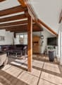 Bright living room with wooden beams, large windows, a dark brown sectional sofa, a coffee table, a leather armchair, a TV, and a wooden cabinet.