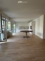 Open-plan space with light wooden flooring, white walls, and a single round table near the kitchen area. Large windows and sliding doors provide natural light and access to the outdoors.
