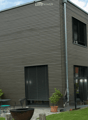Modern house exterior with grey wooden siding, large windows, and a well-maintained garden area featuring potted plants, outdoor furniture, and a fire pit.
