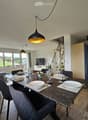 Open-plan living and dining area with a grey sofa, a dark dining table set for six, and a TV on a white stand. The space features wooden flooring, large windows with grey curtains, and modern pendant lights.