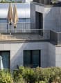 Modern building with grey facade, large windows, and balconies featuring metal railings and closed umbrellas.
