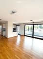 Bright, empty room with wooden flooring, white walls, and large sliding glass doors leading to a balcony. Minimalist design with recessed lighting and a small kitchen area visible in the background.