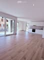 Bright, unfurnished living space with wooden flooring, white walls, recessed lighting, and a large sliding glass door leading to a balcony. Adjacent kitchen area with white cabinets and stainless steel appliances is visible.