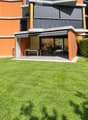 Modern house with orange and grey facade, large lawn, patio with outdoor furniture, and covered entrance area.