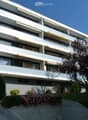 Modern multi-story apartment building with balconies, awnings, and surrounding greenery.