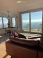 Bright living room with large windows offering a scenic view, featuring a brown leather sofa, throw pillows, a wooden coffee table, and a hanging pendant light.