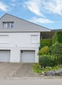 Modern two-story house with white exterior, grey garage doors, and a sloped roof. The house is surrounded by greenery and has a paved driveway leading to the garage.