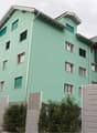 A modern residential building with three floors and several windows. The walls are painted green, and the roof has a triangular shape. The exterior is adorned with multiple white railings on the balcony, and there are bushes and trees on the sides. The bu