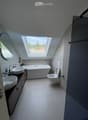Modern bathroom with a large skylight, white bathtub, two sinks, toilet, and a round mirror.